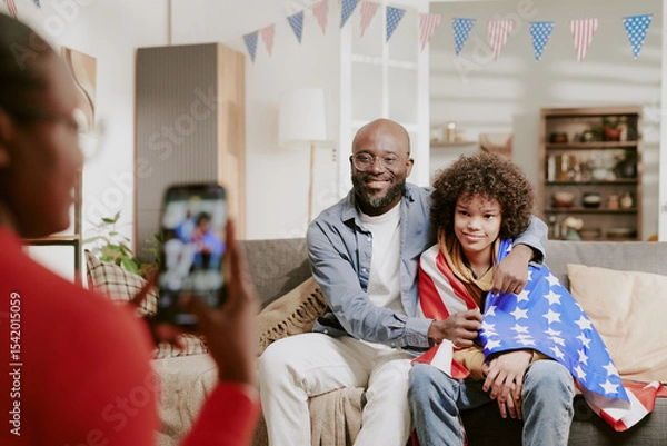 Obraz African American father sitting on sofa embracing teenager wrapped in American flag while woman photographing them with smartphone during family gathering in living room