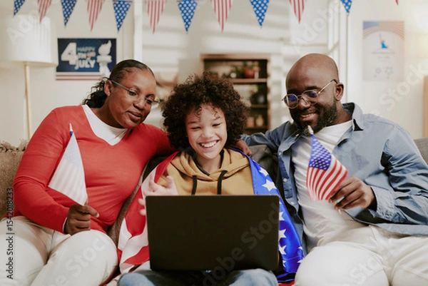 Obraz Family sitting together on sofa smiling and holding American flags while using laptop