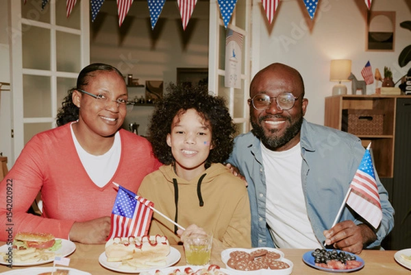 Obraz Portrait of Black woman, Black man, and child smiling and sitting together at table holding American flags, celebrating patriotic holiday with festive food and decorations