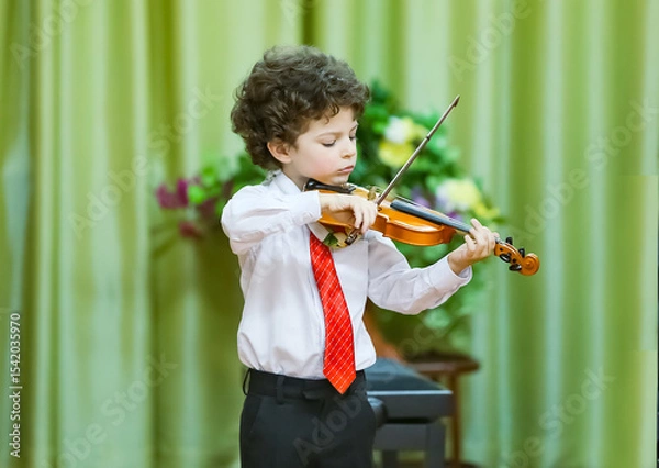 Fototapeta An eight-year-old boy plays the violin at a school concert. He is wearing a white shirt, dark trousers and a tie. He is standing on the stage, holding a violin and a bow, a background curtain