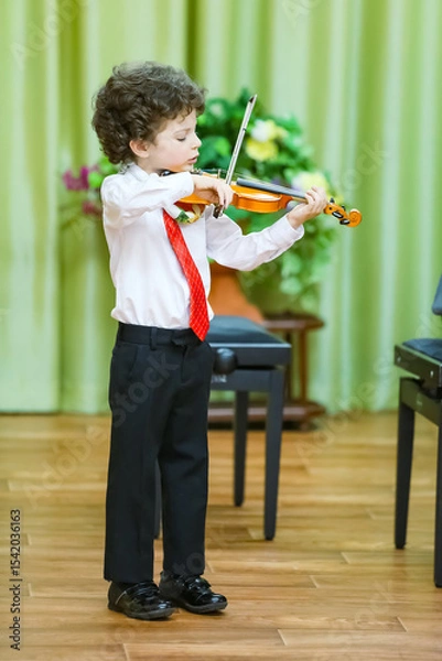 Fototapeta An eight-year-old boy plays the violin at a school concert. He is wearing a white shirt, dark trousers and a tie. He is standing on the stage, holding a violin and a bow, a background curtain
