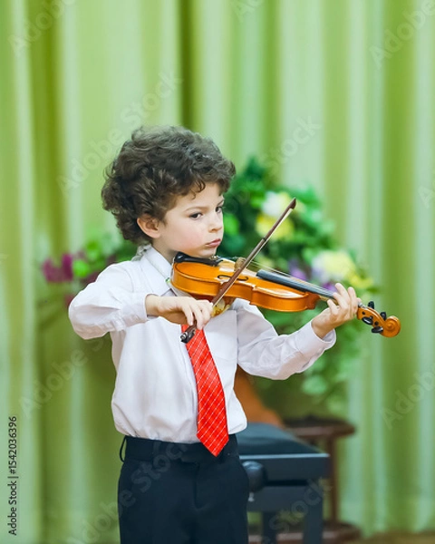 Fototapeta An eight-year-old boy plays the violin at a school concert. He is wearing a white shirt, dark trousers and a tie. He is standing on the stage, holding a violin and a bow, a background curtain