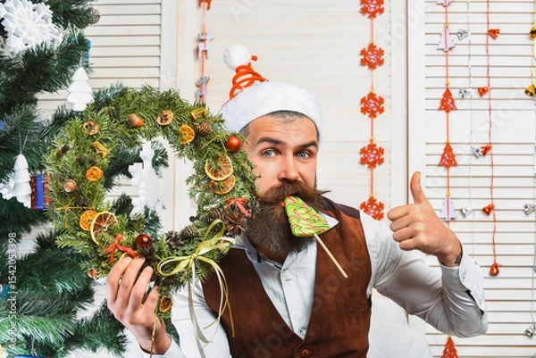 Obraz Xmas decoration for home. Christmas man eat candy. Bearded man at Christmas. New Year holiday. Man with moustache hold Xmas wreath. Happy New Year. Christmas wreath decoration and treats. Thumb up