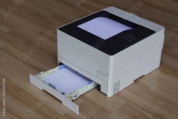 Fototapeta A black and white laser printer sitting on a wooden floor. A stack of white paper is visible in the main paper output tray on top of the printer and the lower paper input tray is pulled out.