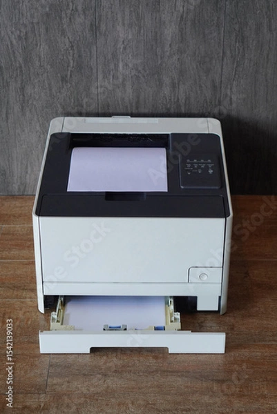 Fototapeta top view  black and white laser printer sitting on a wooden floor. A stack of white paper is visible in the main paper output tray on top of the printer and the lower paper input tray is pulled out.