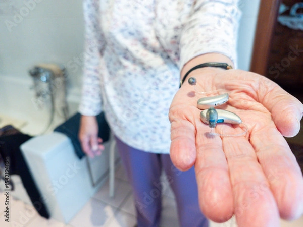 Fototapeta Elderly person holding a pair hearing aids in hand. assistive hearing technology in real-life setting, highlighting accessibility, senior healthcare, hearing loss. Bathroom, emphasizing daily use and 