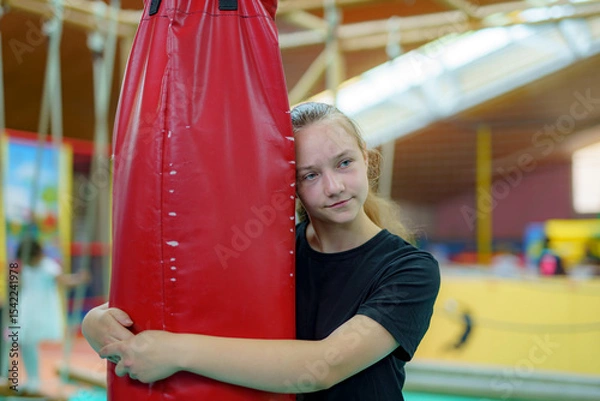 Obraz Young girl embraces a red punching bag while practicing in a gym during a training session