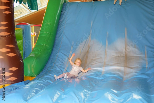 Fototapeta Child joyfully slides down a large inflatable slide in a play area filled with colorful structures and toys