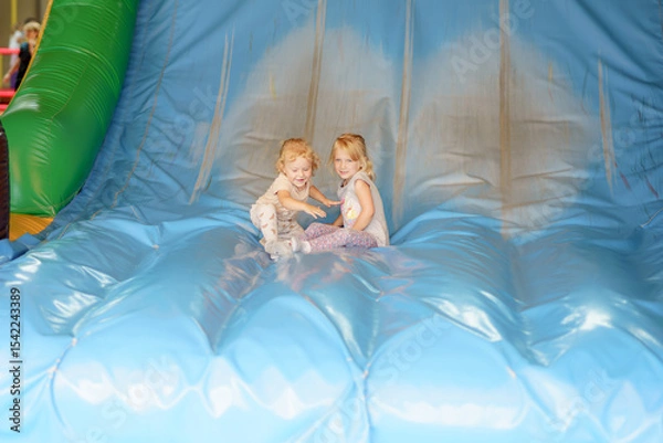 Obraz Two young children enjoying themselves on a large inflatable slide during a fun event at an indoor play center