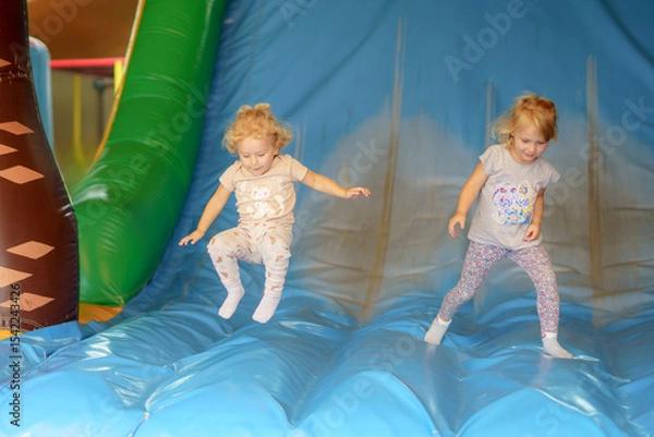 Obraz Two young girls joyfully jumping on a colorful inflatable slide at a play center during a sunny afternoon