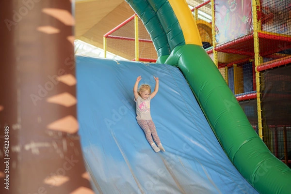 Fototapeta Child joyfully sliding down a colorful inflatable slide in an indoor play area during afternoon playtime