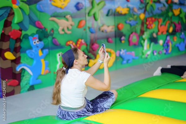 Obraz Woman enjoying smartphone while sitting on a colorful soft surface in a play area with vibrant wall decorations