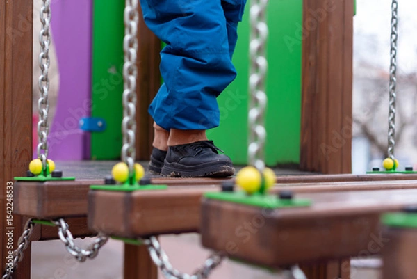 Fototapeta Child walking on playground bridge with chains and colorful wooden elements.