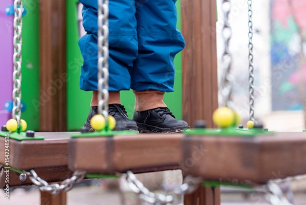 Fototapeta Child walking on playground bridge with chains and colorful wooden elements.