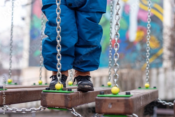 Fototapeta Child walking on playground bridge with chains and colorful wooden elements.