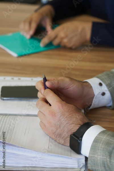 Obraz Close up of a businessman’s hands holding a pen over an open notebook during a meeting, ideal for illustrating corporate note-taking, planning and teamwork. Photo