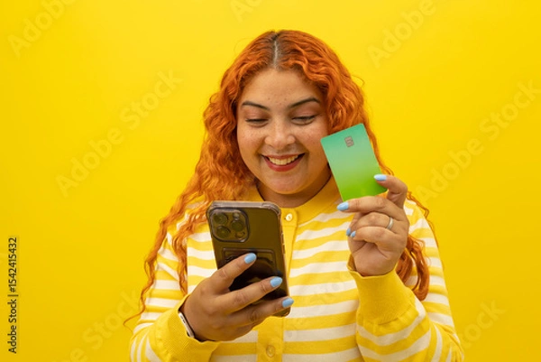 Fototapeta Cheerful young woman with curly red hair holding a card while shopping from her cell phone on a yellow background, concept of joy when shopping from cell phone