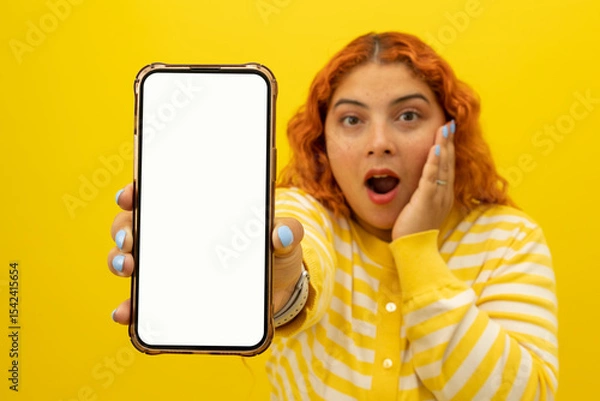 Fototapeta Young freckled woman with curly red hair with her hand on her face making a surprised expression showing her cell phone with a blank screen on a yellow background with copyspace