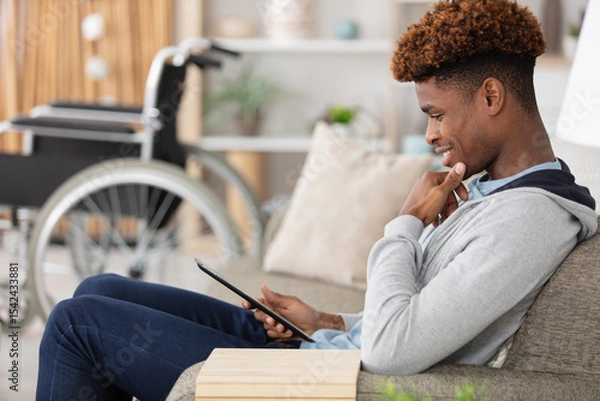 Obraz man relaxing on sofa with tablet near wheelchair