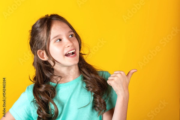 Obraz Portrait of cute young caucasian european blonde girl with opened mouth pointing thumb finger to the side side on yellow background. Studio shot