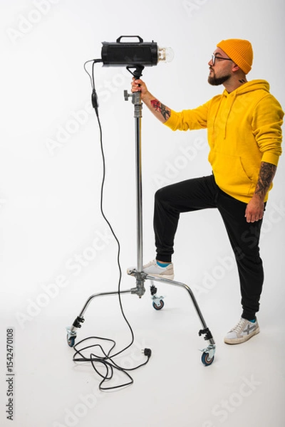 Obraz Smiling man in yellow hoodie holding professional lighting equipment in studio. Behind-the-scenes concept with cheerful crew member during photo or video shoot setup