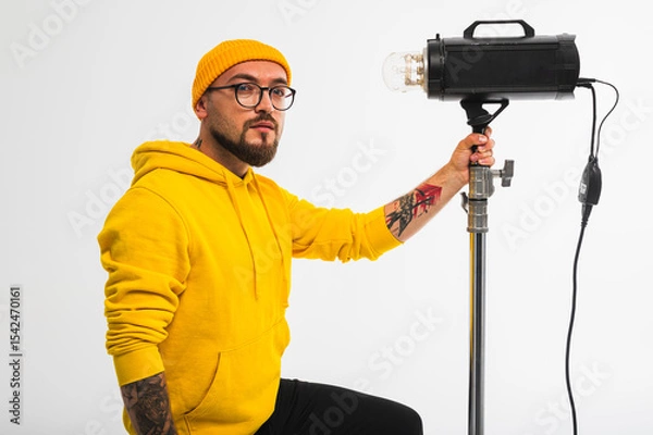 Obraz Smiling man in yellow hoodie holding professional lighting equipment in studio. Behind-the-scenes concept with cheerful crew member during photo or video shoot setup