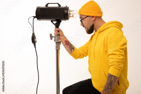 Obraz Smiling man in yellow hoodie holding professional lighting equipment in studio. Behind-the-scenes concept with cheerful crew member during photo or video shoot setup