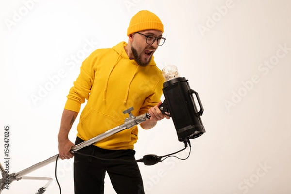 Obraz Smiling man in yellow hoodie holding professional lighting equipment in studio. Behind-the-scenes concept with cheerful crew member during photo or video shoot setup