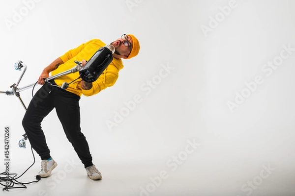 Obraz Smiling man in yellow hoodie holding professional lighting equipment in studio. Behind-the-scenes concept with cheerful crew member during photo or video shoot setup
