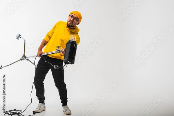 Obraz Smiling man in yellow hoodie holding professional lighting equipment in studio. Behind-the-scenes concept with cheerful crew member during photo or video shoot setup