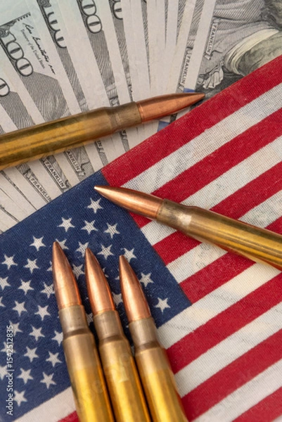 Obraz Close-up of firearm ammunition lying on an American flag with blurred dollar notes in the background. The image symbolises the themes of war, security, power and financial influence in the USA.