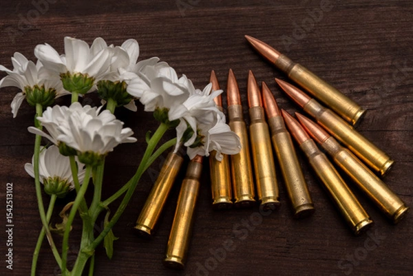 Obraz A bouquet of white chrysanthemums lies next to rifle cartridges on a dark wooden surface. The image symbolises the contrast between peace and war, life and death, or the memory of the fallen.