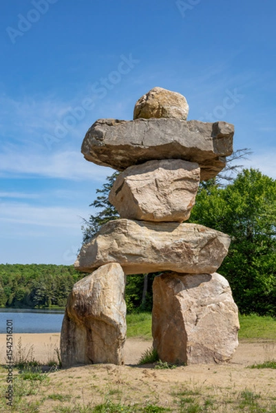 Fototapeta A towering Inukshuk stands proudly by the serene shores of Clear Lake, set against a backdrop of lush greenery and a clear blue sky.