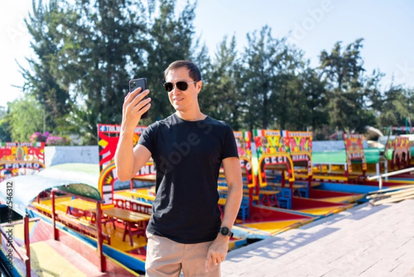 Fototapeta Hispanic Man Using Smartphone in Xochimilco with trajinera boats in the background, Mexico