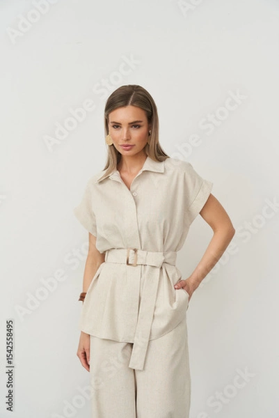 Fototapeta Neutral-toned outfit with a belted top and matching pants, worn by a woman with straight hair and large earrings, standing with one hand in a pocket, on a white backdrop