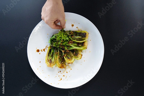 Obraz A hand adds garnishing to freshly prepared bok choy on a white plate in a vibrant kitchen setting.