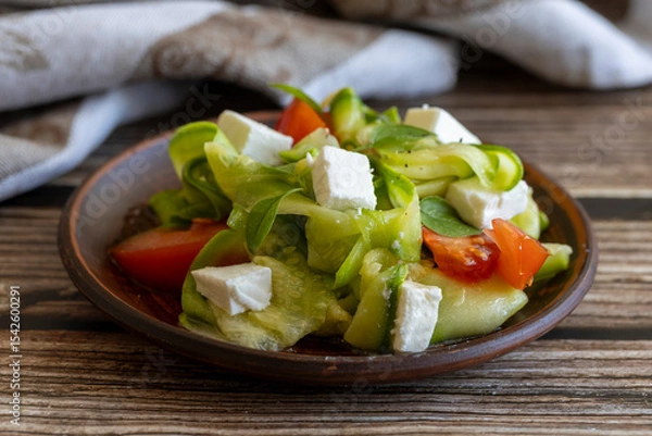 Fototapeta Vegetable salad with raw pickled zucchini, feta cheese and fresh tomatoes on clay plate with cloth napkin on wooden table, side view, close up. Healthy diet.