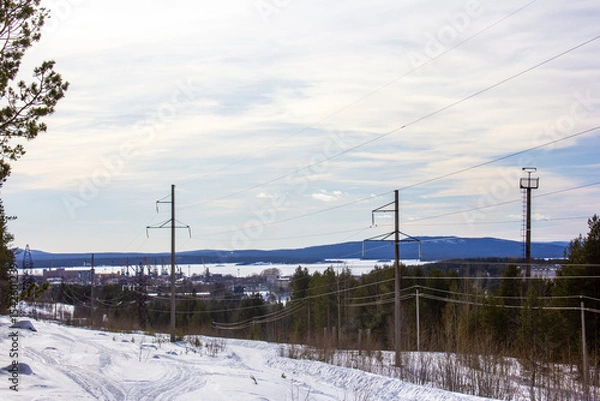 Fototapeta  A snow-covered clearing with power lines stretching across the winter landscape. Tall pylons line up along the edge of a pine forest under a pale blue sky, creating a mix of nature and infrastructure