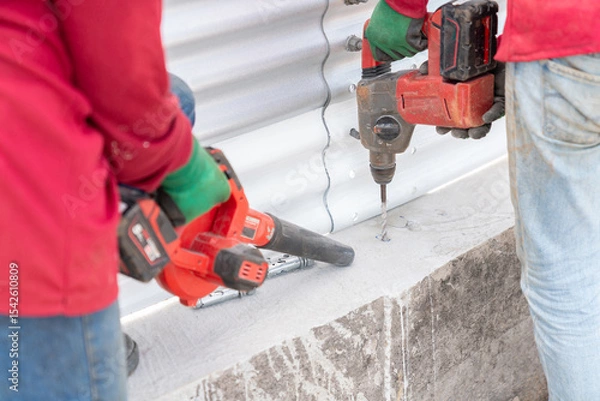 Fototapeta Two workers are using a cordless drill to drill into the concrete floor, while one of them is using a cordless blower to remove dust and debris.