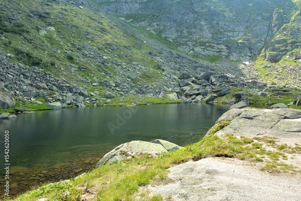 Obraz A small calm lake at the foot of a steep rocky slope of a high cliff on a sunny summer morning.