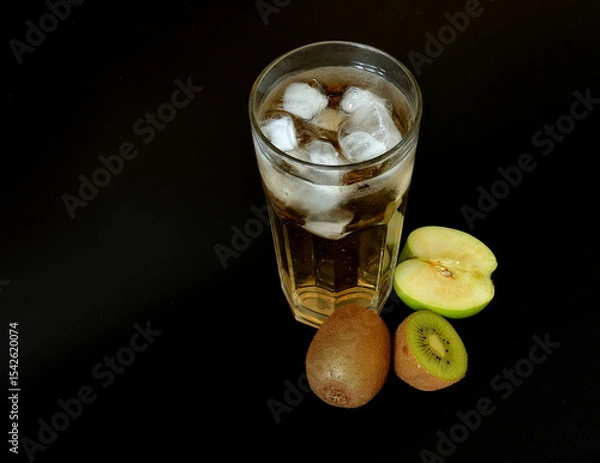 Obraz Tropical fruit juice in a tall glass with ice on a black background, next to it are pieces of ripe kiwi and green apple.