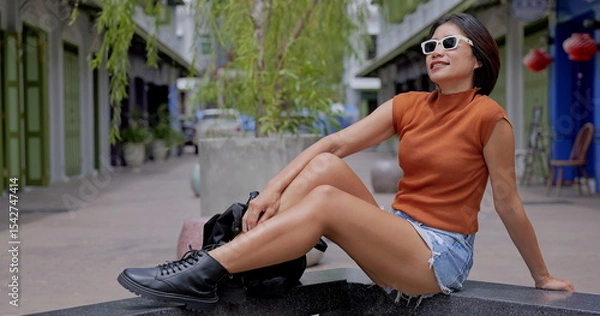 Obraz Asian woman wearing sleeveless turtleneck top, distressed denim shorts and black combat boots enjoys the sun on a sleek stone bench in a charming urban alleyway