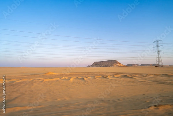 Obraz The vast Aswan desert landscape at dawn, showcasing unique rock formations and a prominent power line tower under a clear sky, along the route from Aswan to Abu Simbel.