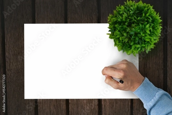 Fototapeta Flat design is showing hand holding black pen over blank paper on wooden tabletop with plant