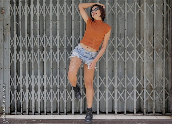 Obraz Pretty, chic Asian woman poses stylishly against a metal gate, in a rust crop top, distressed denim shorts, and black combat boots, looking cute with an urban edge