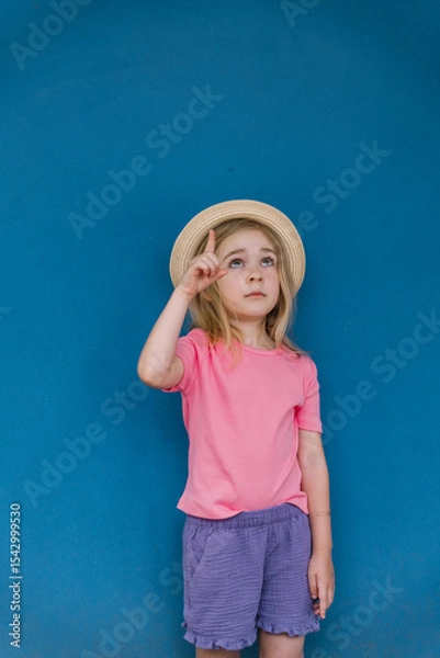 Fototapeta Young girl with a straw hat pointing upwards against a blue wall in bright summer attire