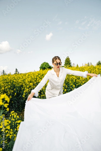 Fototapeta Model poses in a bright yellow flower field while holding a flowing white fabric on a sunny day
