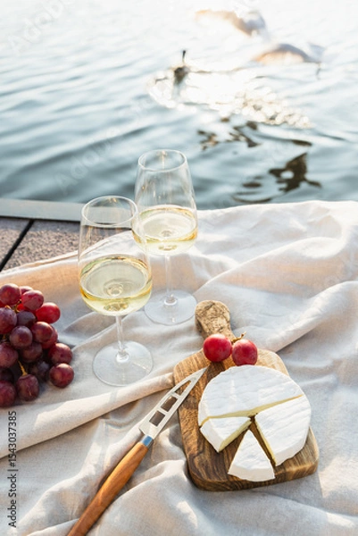 Fototapeta Two glasses of white wine with cheese and grapes on a pier at sunset.