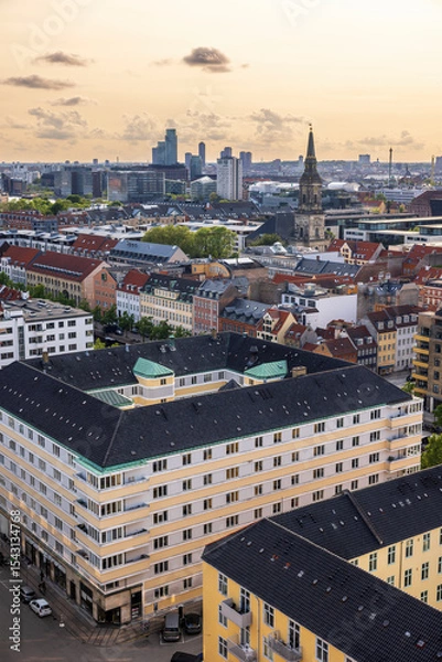Fototapeta Panoramic view of Copenhagen taken from the top of the Church of Our Saviour, showing a mix of historical rooftops, modern buildings, and city landmarks under a warm evening sky.