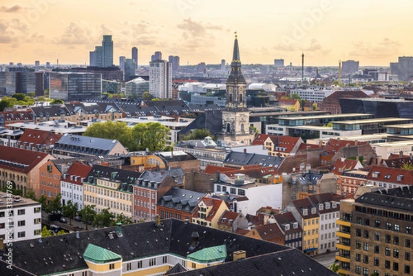 Fototapeta Panoramic view of Copenhagen taken from the top of the Church of Our Saviour, showing a mix of historical rooftops, modern buildings, and city landmarks under a warm evening sky.